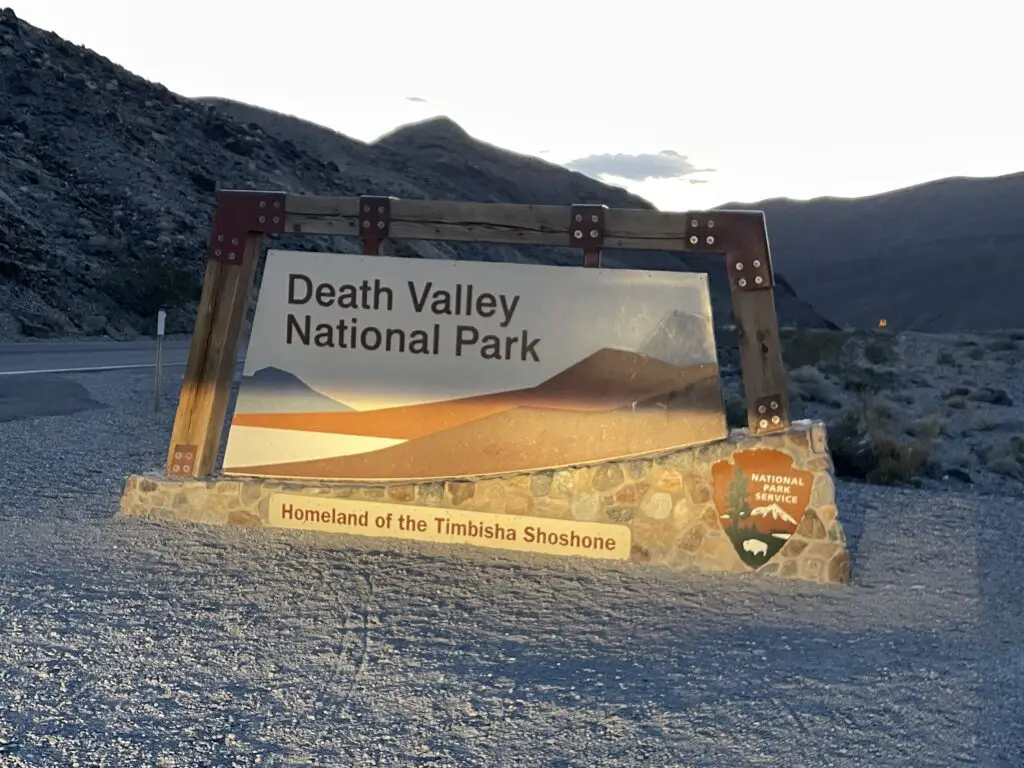 National park entrance sign with rocky, desert mountains and a sunset in the background. The sign reads Death Valley National Park. 