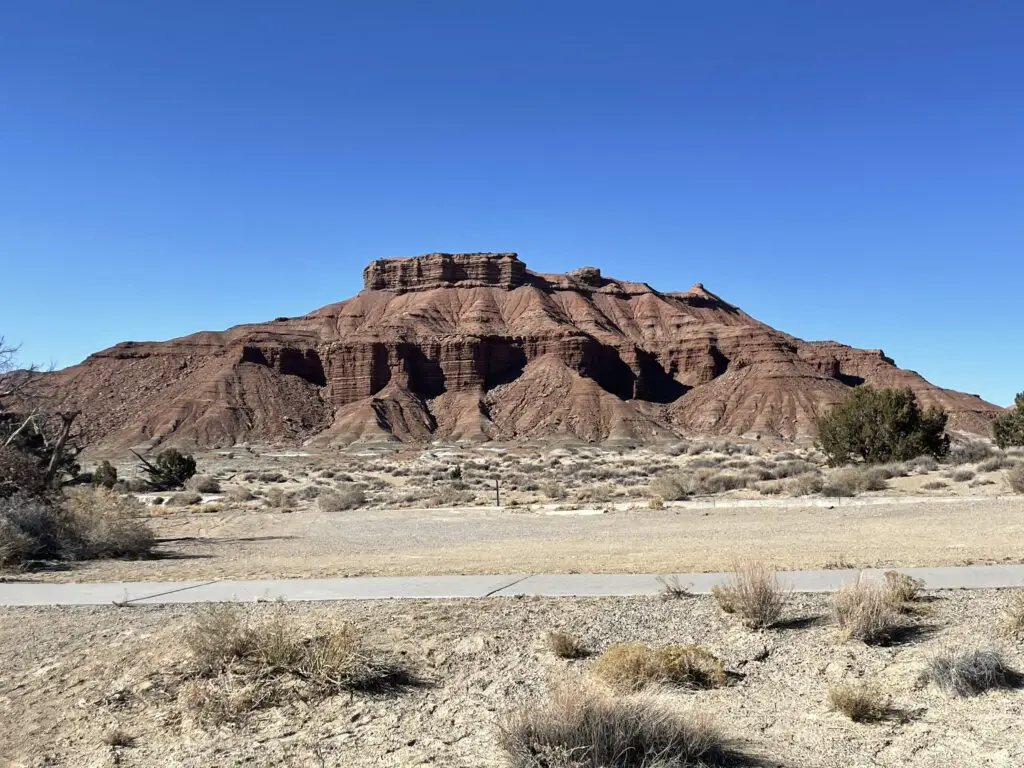 A red-rock mountain rises from a flat, yellow, scrub brush-covered, desert landscape.