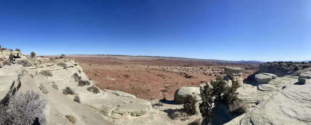 Panoramic picture of grey rock in the foreground overlooking a flat, red-rock plain, all under a clear, blue sky