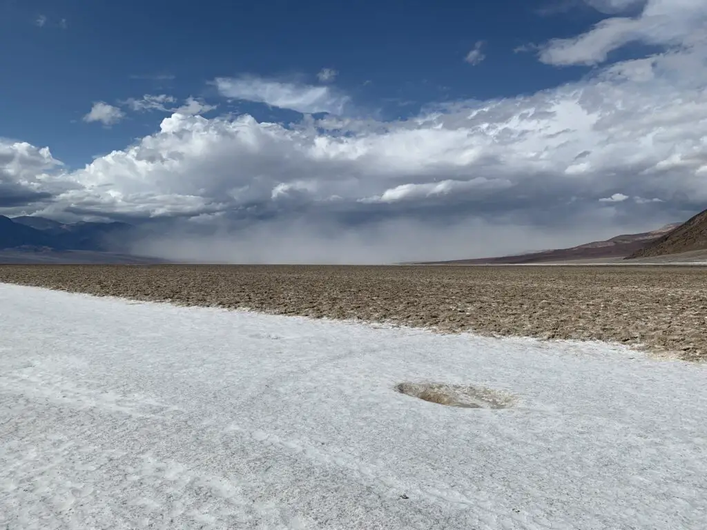 A rocky, salt-flat spreads to the horizon, surrounded by rocky mountains. A dusty cloud fills the valley.