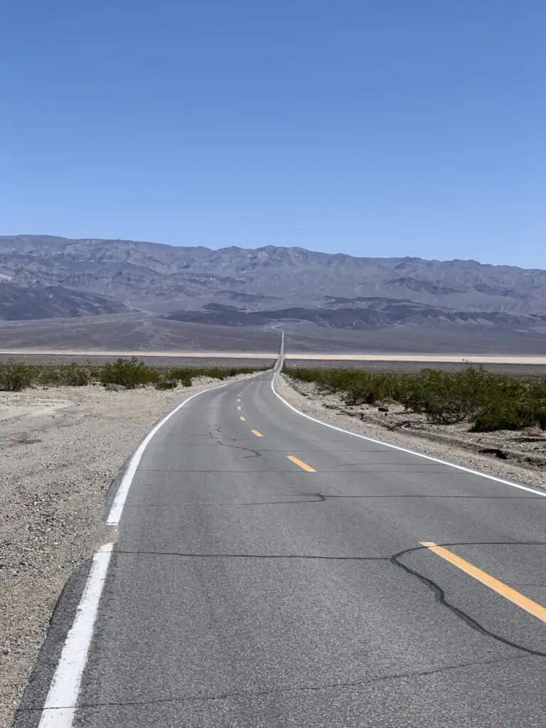 Empty, two-lane road stretches across a rocky, flat desert (with scrub brush) to a rocky mountain towering far in the distance