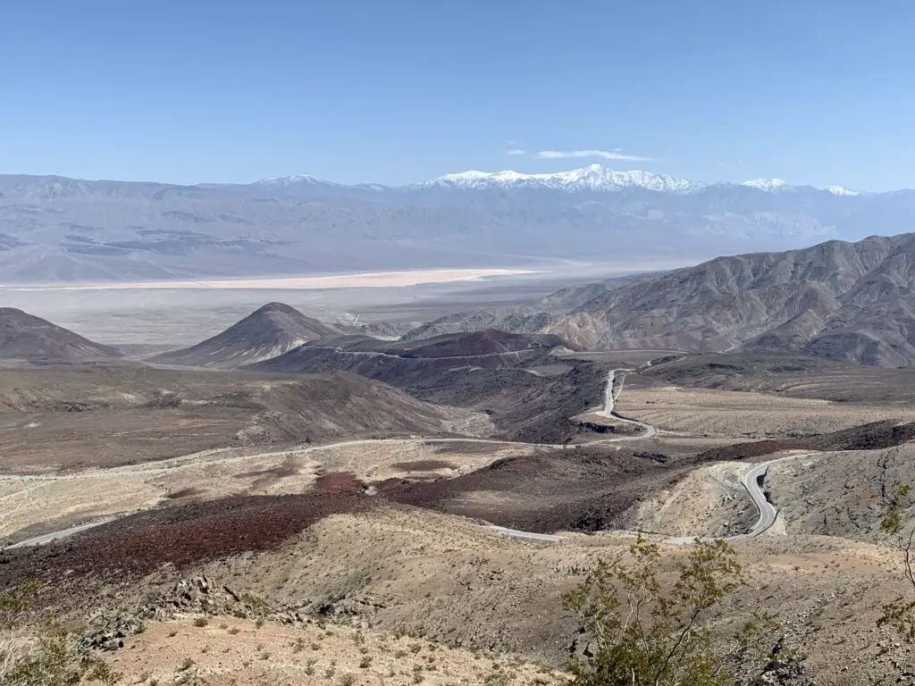 View down a rocky hillside with red, brown, and tan gravel. A grey, flat valley is in the background backed by a rocky, snow-capped mountain. 