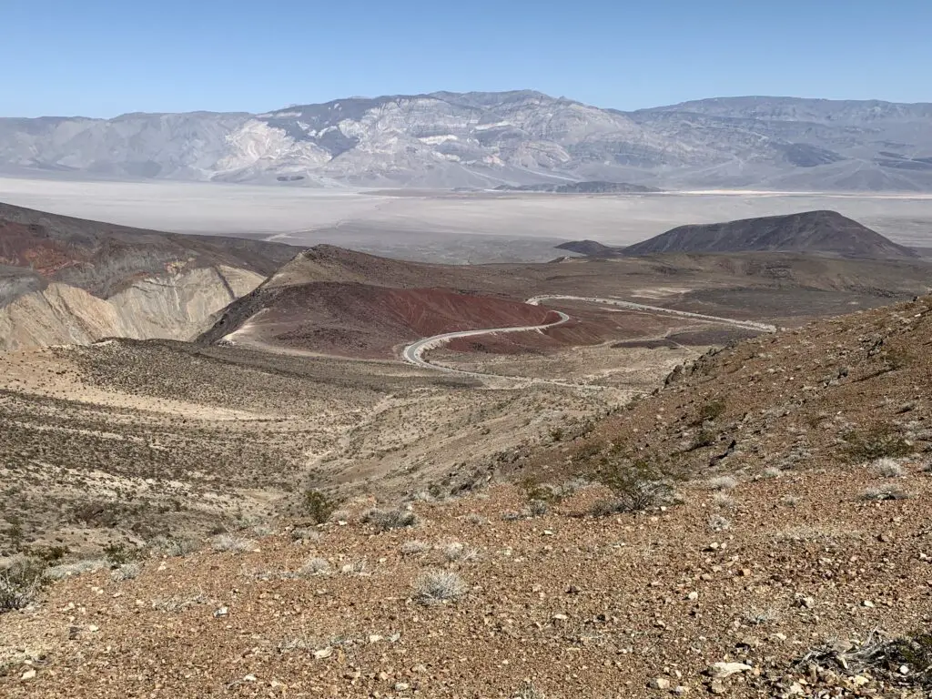 View down a rocky hillside traversed by a road with red, brown, and tan gravel. A grey, flat valley is in the background backed by a rocky mountain. 