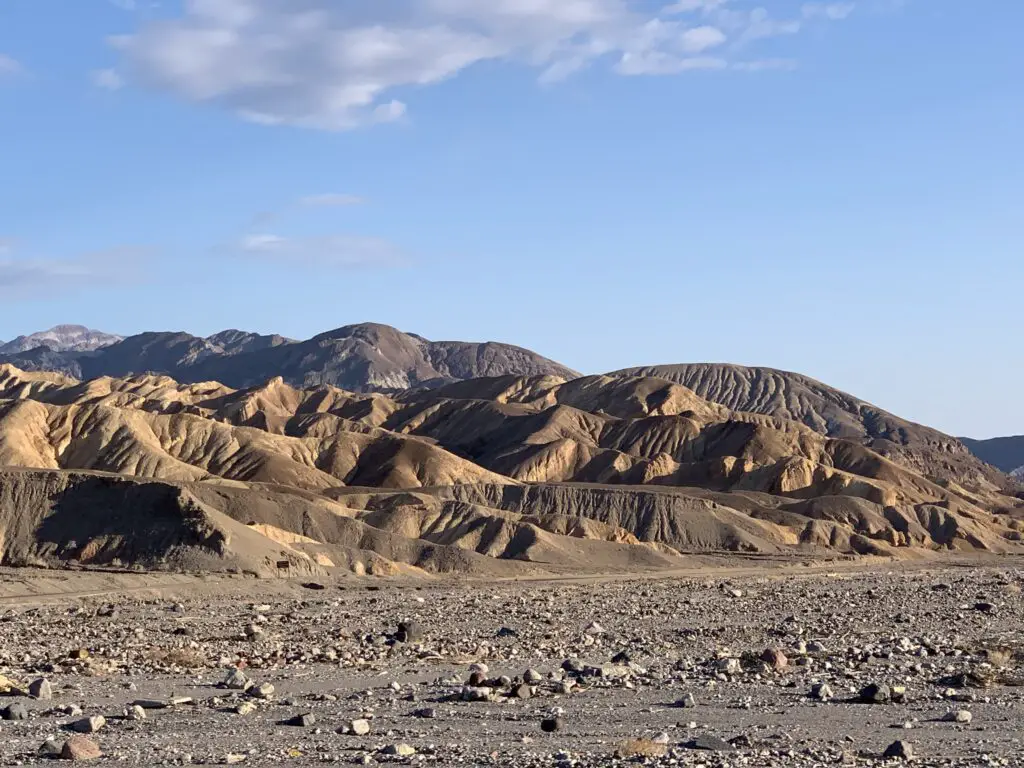 A flat, gravel plain in the foreground with brown, gravel hills rising in the background under a blue sky. 