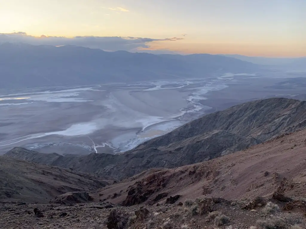 View from above, looks down a rocky and scrub brush-covered cliff to a gravely plain streaked with salt stains and water puddles. An orange sunset and clouds cover dark mountains in the distance.