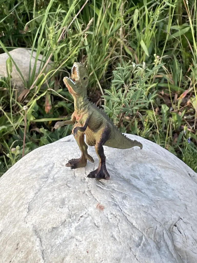 A toy dinosaur with it's head back roaring sits on top of a rock with tall grass surrounding it