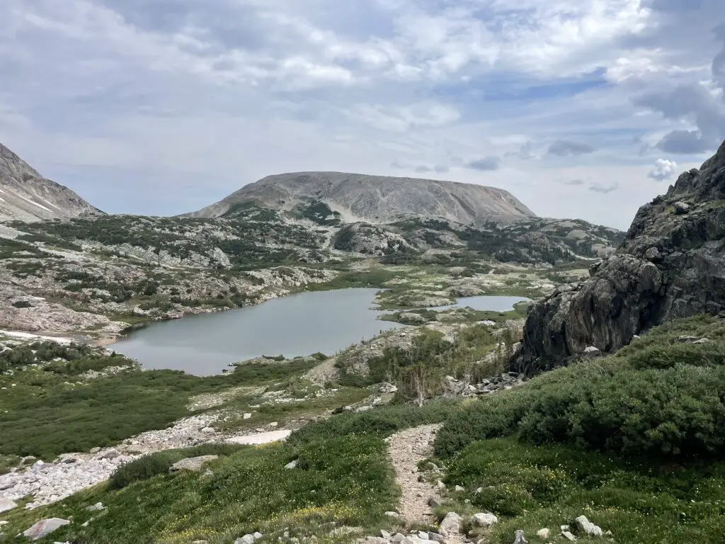 Grey, rocky mountains rise behind two small lakes and flat scrubland. A rocky trail rises up a grassy hill in the foreground.