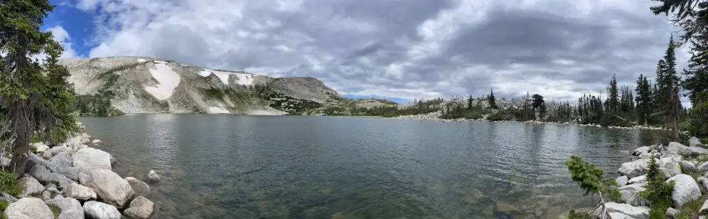 A panoramic of a large, alpine lake with gray, stony mountains spotted with snow looming in the background.