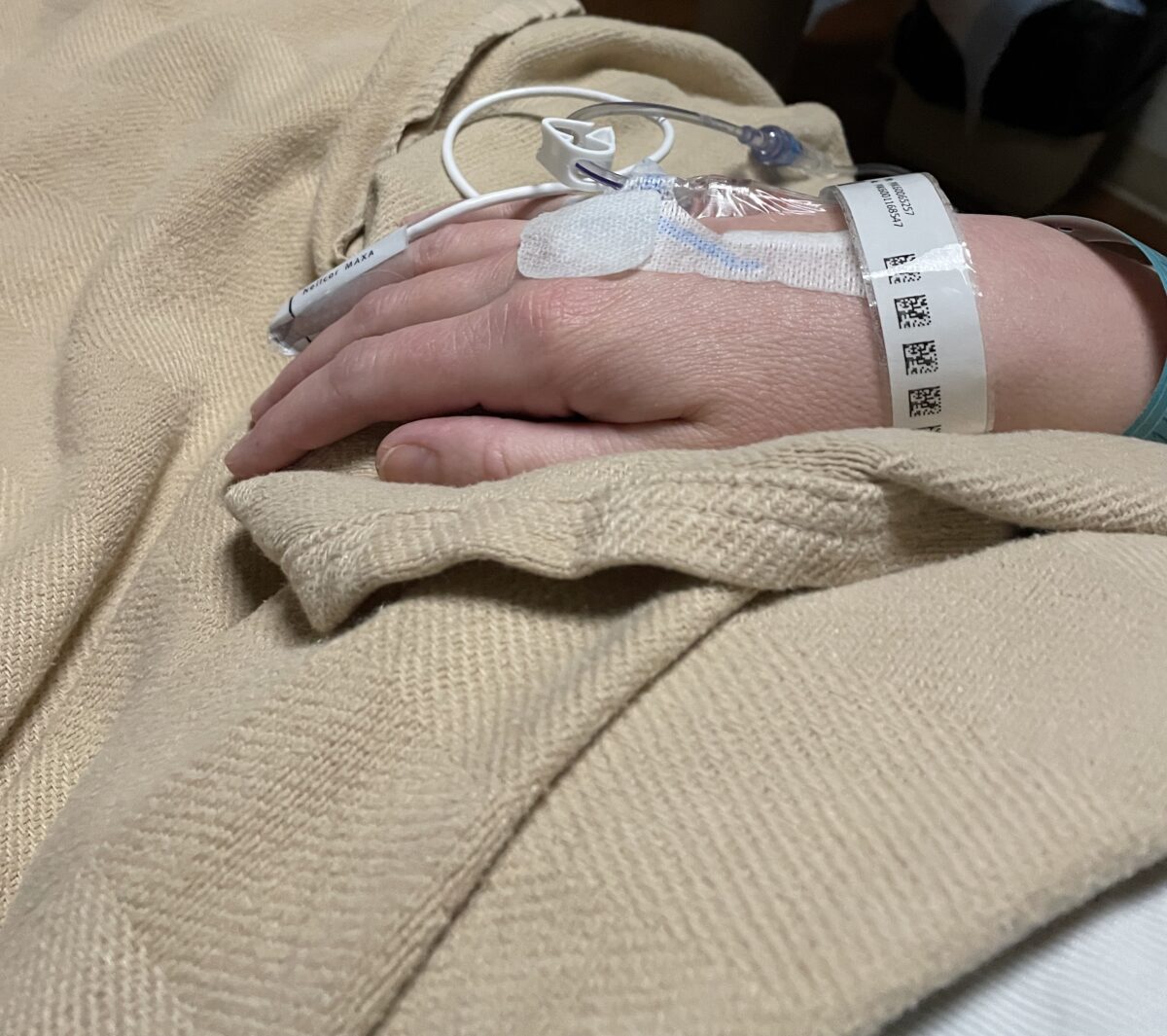 A hand with an IV sits on a hospital bed