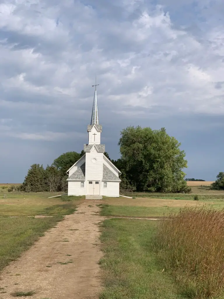 A white church stands amongst some trees in a field with a dirt path leading to it