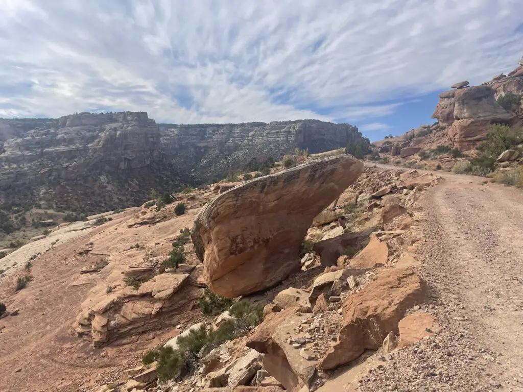 A large boulder in the shape of an alien head and face sit alongside a dirt trail with rocky canyon walls in the background. 