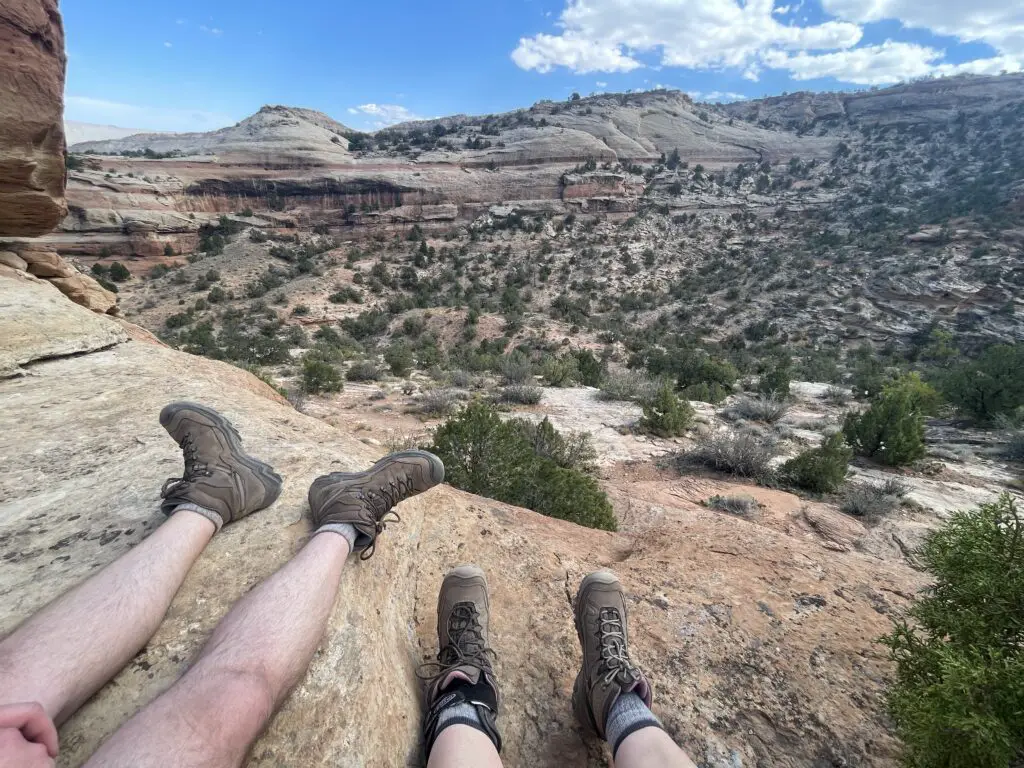 Two pairs of hikers legs and feet-in-boots sitting on a stone overlooking a dry canyon filled with green scrub brush under a blue sky. 