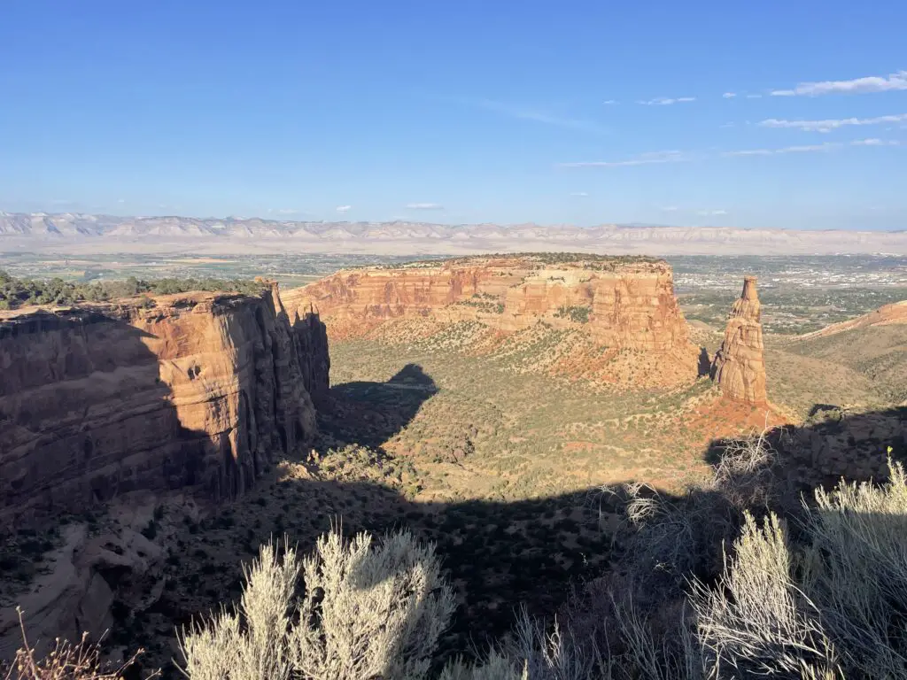 Red rock formations rise from a scrub brush-covered canyon floor, all under a clear, blue sky