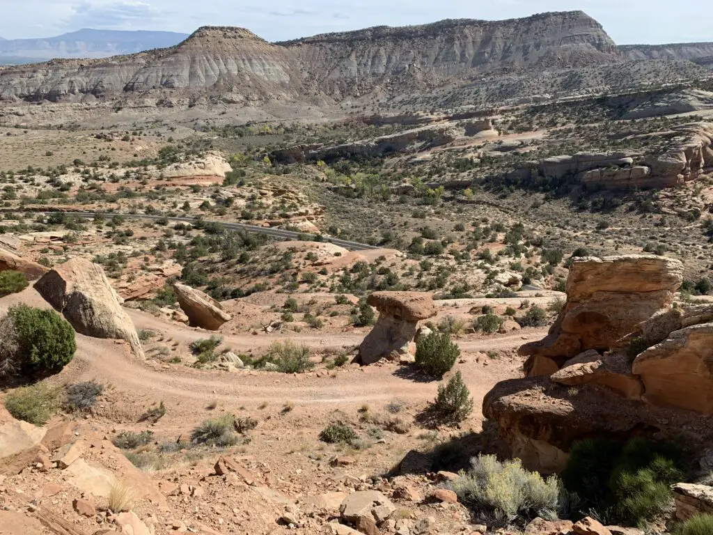 Rocky, gray cliffs in the background overlook a red-gravel and green scrub brush-filled canyon. 