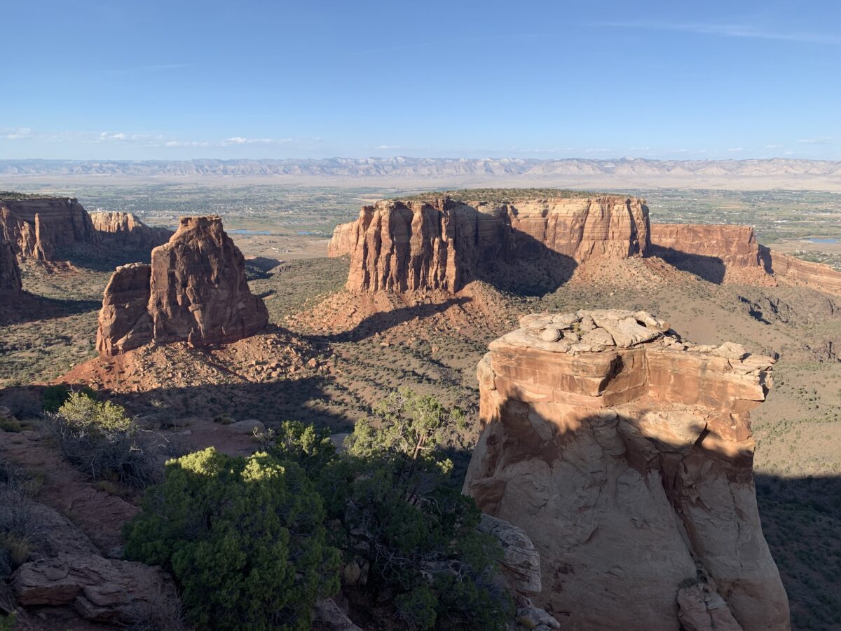 Multiple red-rock monuments surrounded by green scrub brush in canyon country; all under a clear, blue sky.