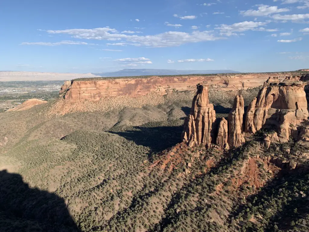 Scrub-brush covered canyon floor with red rock walls rising on the sides; brown, rocky cliffs in the background all under a blue sky with some white, puffy clouds. Rock monoliths also rise from the canyon floor.