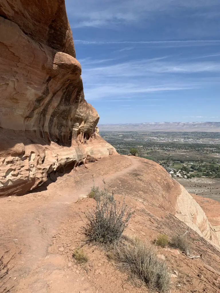 A dirt trail traverses a narrow, rock ledge with rock walls on one side and steep drop-off on the other,