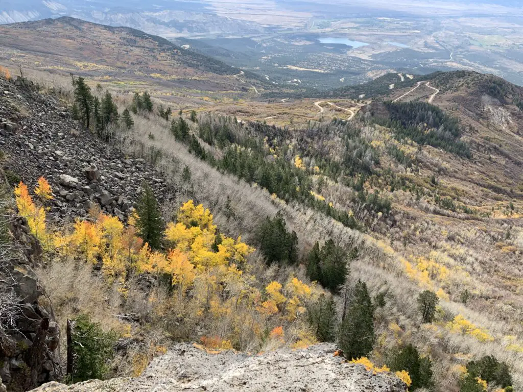 View down a mountain slope with yellow-leafed and dark, pine green trees. A dirt road snakes along farther down the hill and small lakes can be seen in the far distance. 