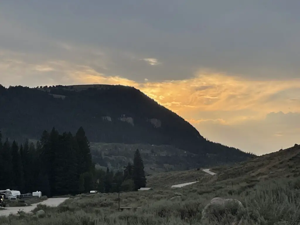 Sunset view of gray clouds over a tree-covered mountain in the background with a small campground and campers in the foreground--the whole picture looks a little hazy due to wildfire smoke. 