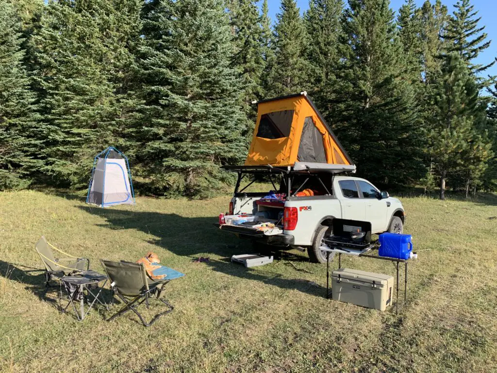 Opened-tent on top of truck with camping equipment and dog scattered about a meadow-area with pine trees in the background