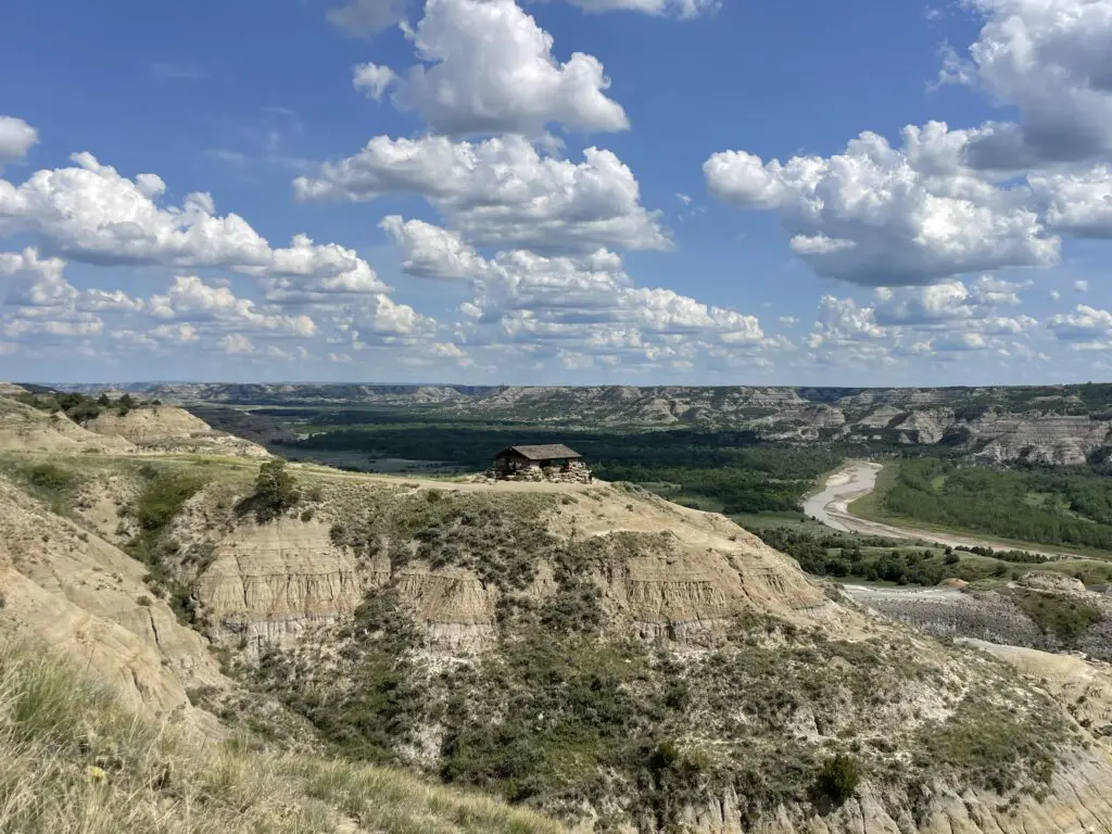Seen from a distance: a large, stone, picnic shelter sits on top of a rocky, tan hill. A green, river valley and other tan, rock and grass-covered hills are in the background all under a blue sky spotted with white, puffy clouds.