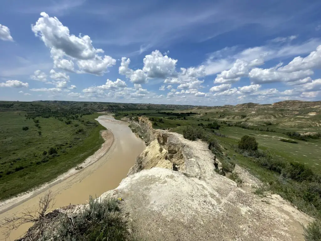 A wide, brown river meanders through a green, grass field with a tan, rocky outcrop on one side, all under a blue sky with puffy, white clouds