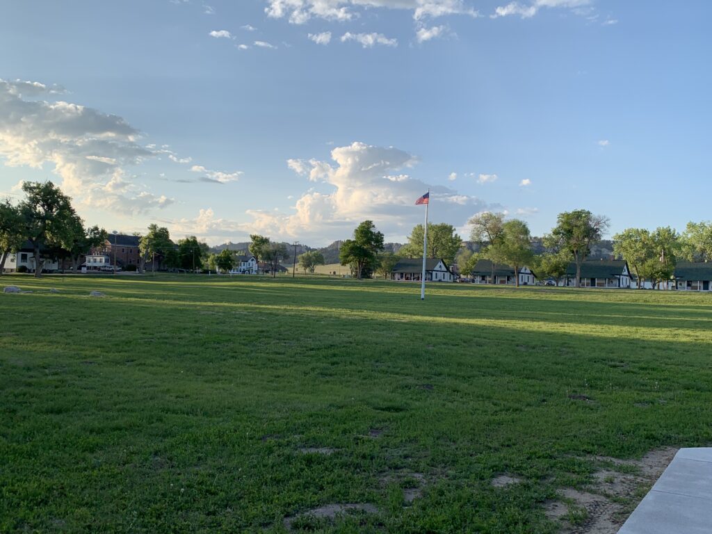 A wide grassy field with an American flag on a flagpole in the center, surrounded by small buildings and trees under a blue sky with scattered clouds. 