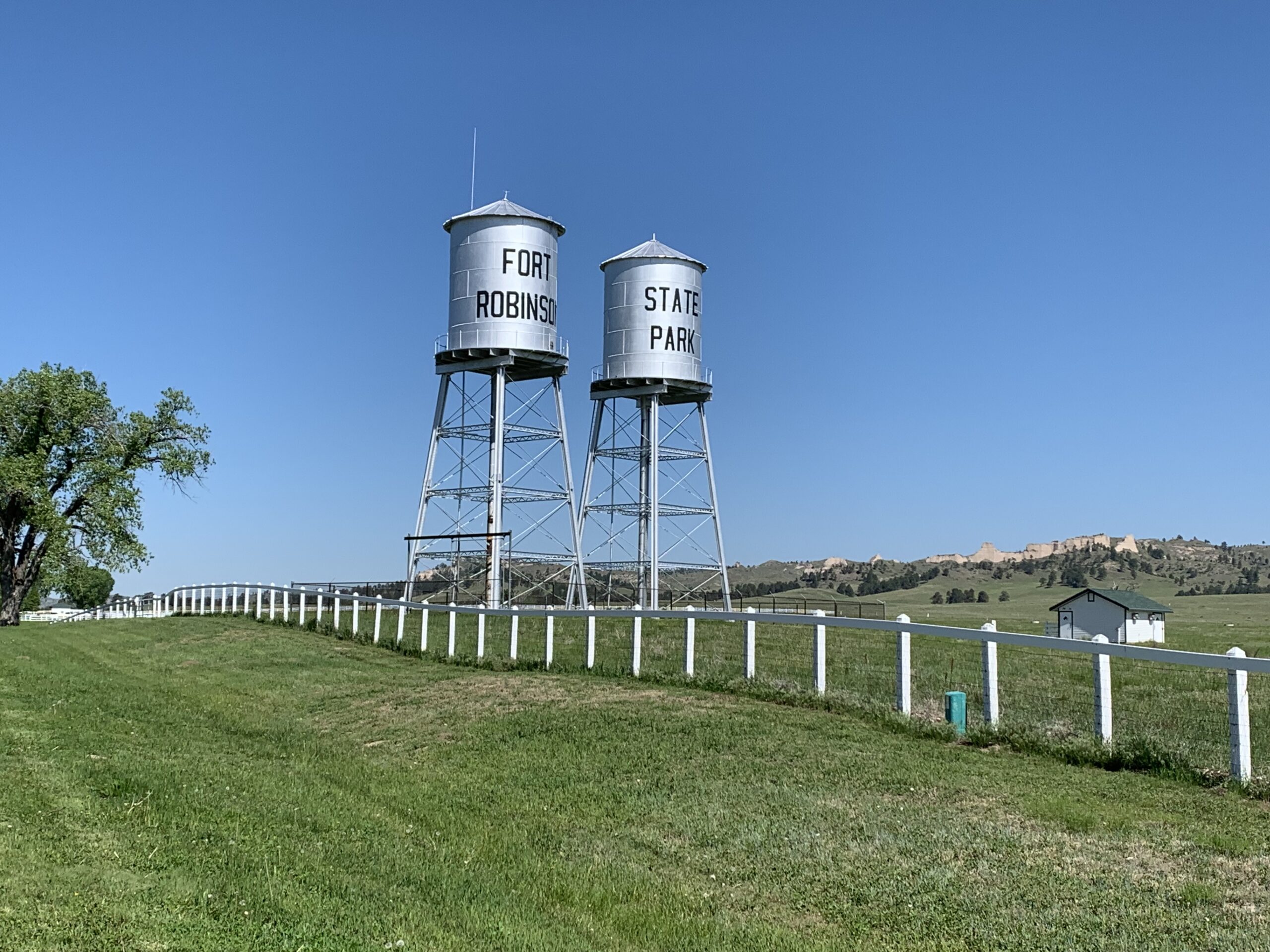Two metal, water towers stand in a green, grass field under a clear, blue sky. The one on the left has "Fort Robinson" and the one on the right has "State Park" written on them.