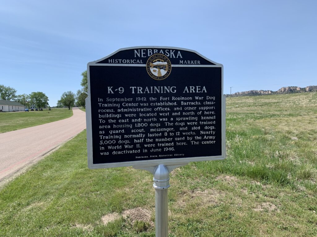 A Nebraska historical marker titled K-9 Training Area stands by a road and grassy field, describing the World War II war dog training center established in 1942 and deactivated in 1946.