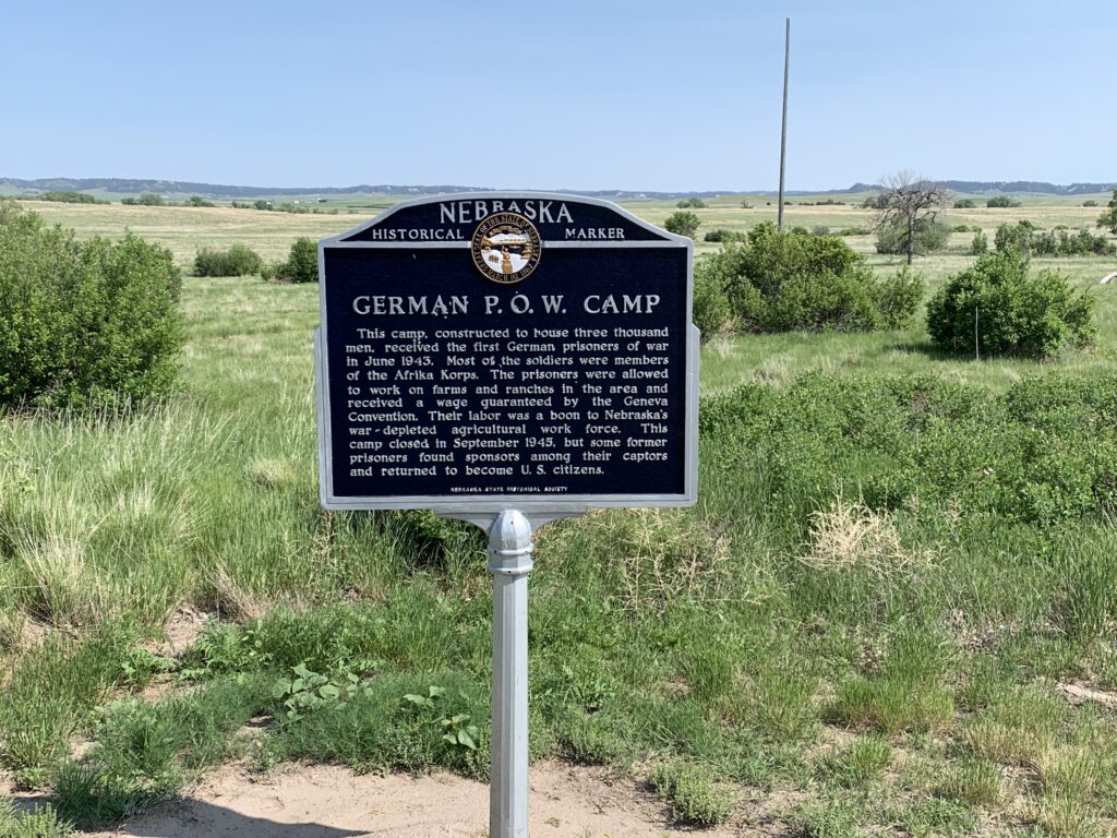 A Nebraska Historical Marker stands in a grassy field. The sign reads “German P.O.W. Camp” and provides historical information about a World War II camp for German prisoners of war in Nebraska. Bushes and open plains are visible behind.