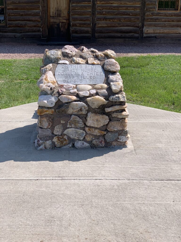 A stone marker reads "On this spot, Crazy Horse Ogallala Chief was killed, Sept 5, 1877". A wood cabin is in the background. 