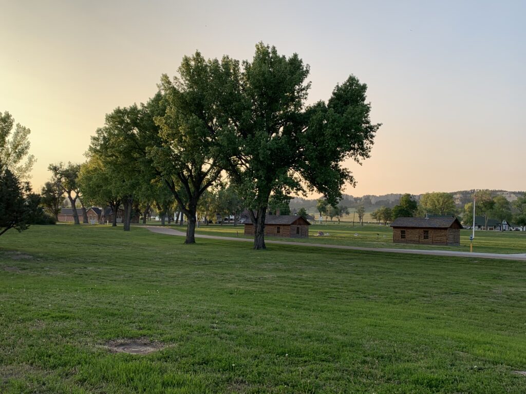 A grassy park at sunset with several tall trees and small wooden cabins spaced throughout the area, under a clear sky. 
