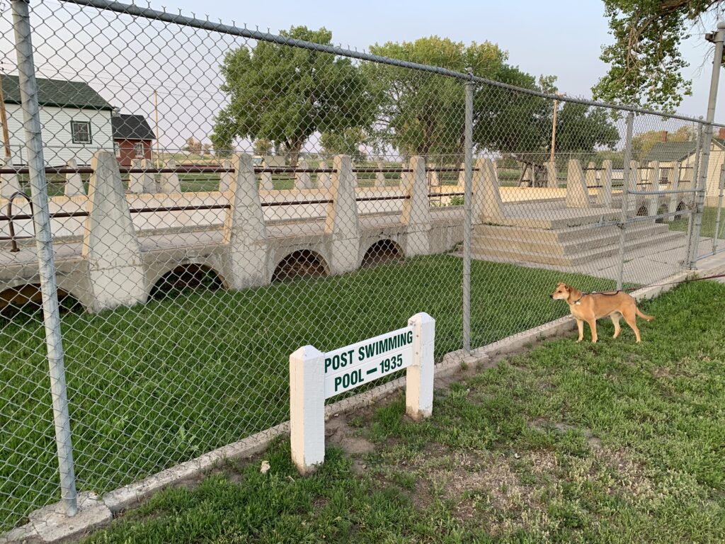 A fenced-off historic pool structure labeled Post Swimming Pool – 1935 stands behind a chain-link fence, with a dog looking through the fence. Trees and houses are visible in the background.