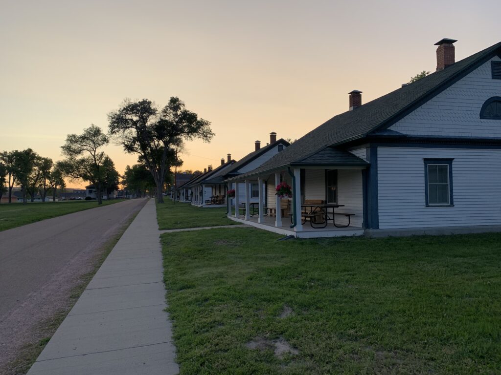 A row of white houses with porches lines a quiet street at sunset, with a sidewalk and grass in the foreground and trees in the background.
