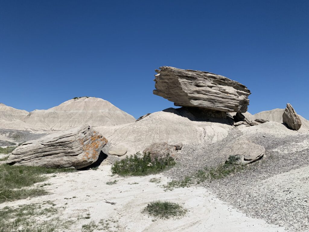 A large, flat boulder is precariously balanced on a mound of pale, rocky ground under a clear blue sky.