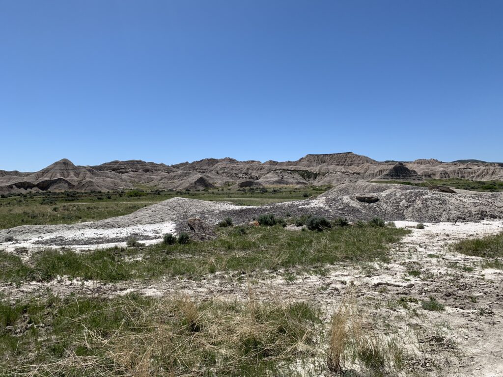 A dry landscape featuring rocky hills under a clear blue sky, with sparse green grass and patches of white, sandy soil in the foreground.