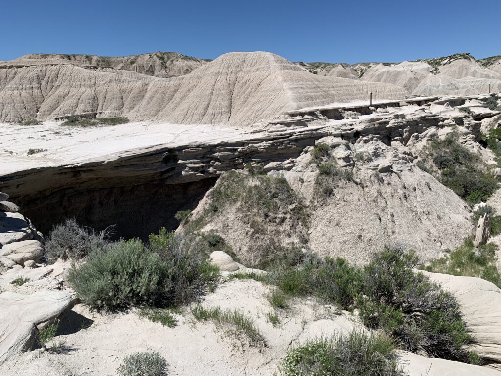 A natural stone bridge spans a deep ravine in a dry, rugged, badlands landscape under a clear blue sky, surrounded by sparse green shrubs and layered rock formations.