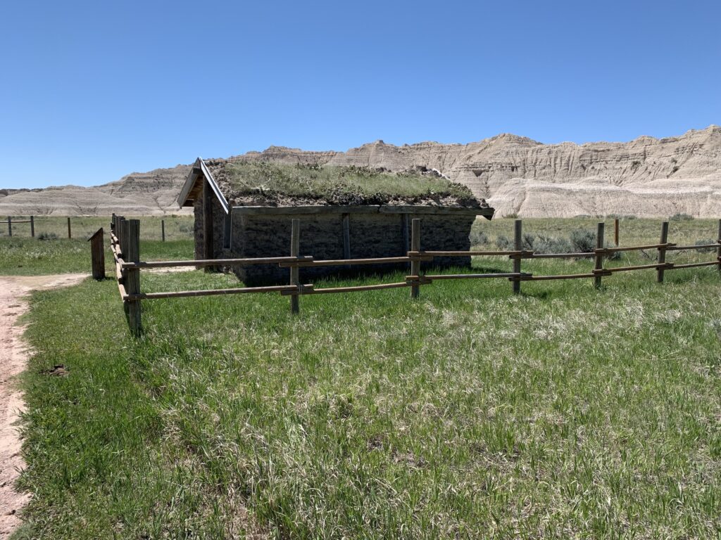 A small log cabin with a sod-covered roof stands behind a wooden fence in a grassy field, with rocky hills and a clear blue sky in the background.