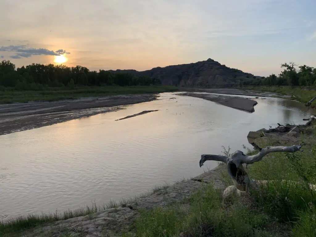 A calm river winds through rocky landscape at sunset, with gentle reflections on the water, grassy banks, a driftwood log in the foreground, and hills silhouetted against a soft, colorful sky.