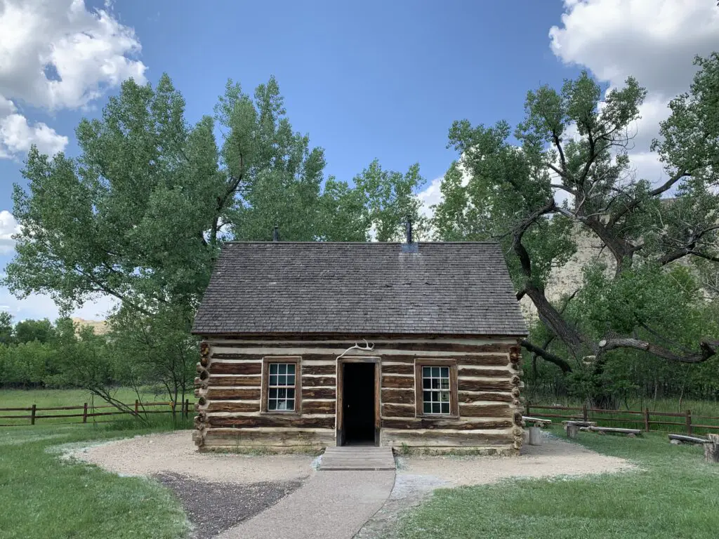 Log cabin with green trees in the background under a blue sky with white clouds