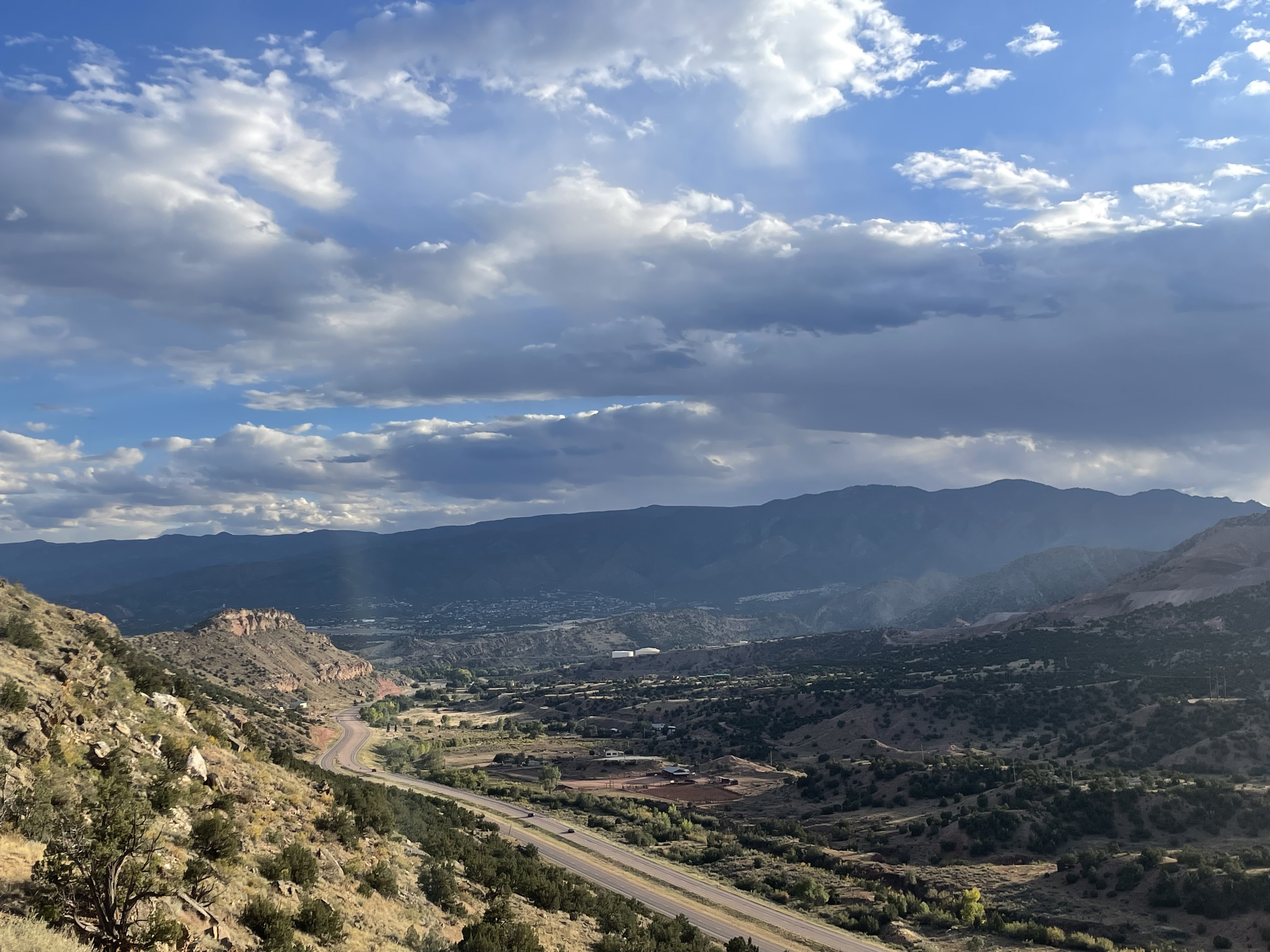 A scenic view of a winding road passing through a valley with hills, scattered vegetation, and mountains in the distance under a partly cloudy blue sky. Sunlight breaks through the clouds, casting light on the landscape.