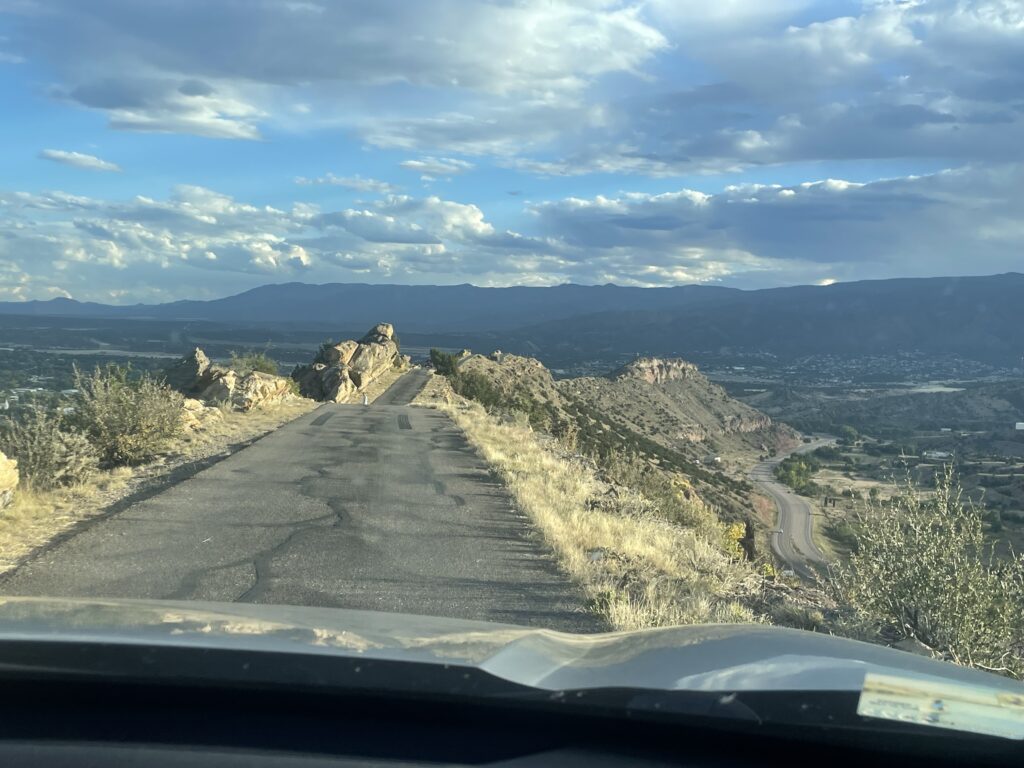 View of a narrow, mountain road with dropoffs on both sides and rocky edges, overlooking a scenic valley and distant mountains under a partly cloudy sky.