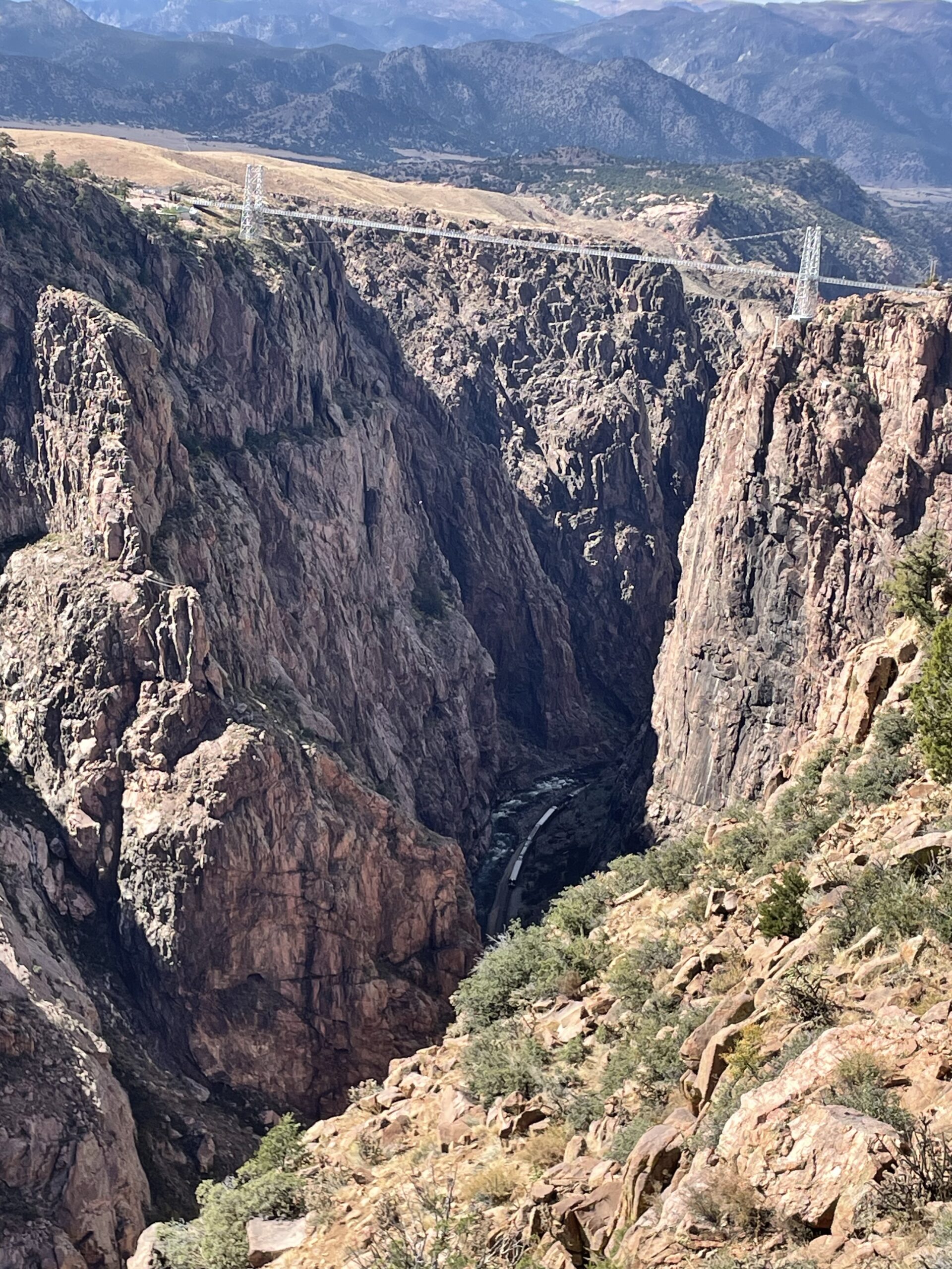 A deep rocky gorge with steep cliffs, spanned by a suspension bridge on the rim. A river flows at the bottom of the canyon, surrounded by rugged terrain and sparse vegetation. Mountains are visible in the distant background.