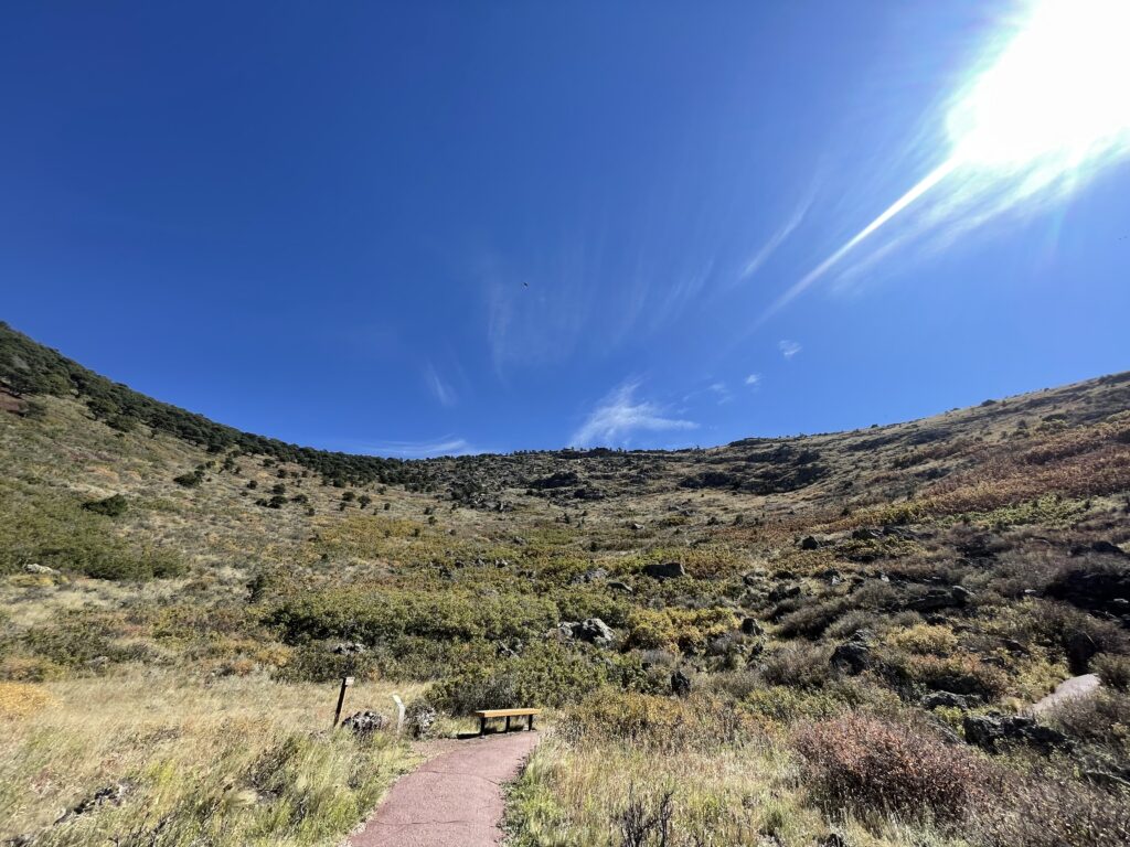 View from the bottom of a crater...a dirt path leads to a wooden bench surrounded by dry grass and shrubs at the base of a sunlit, sloping hillside under a bright blue sky 