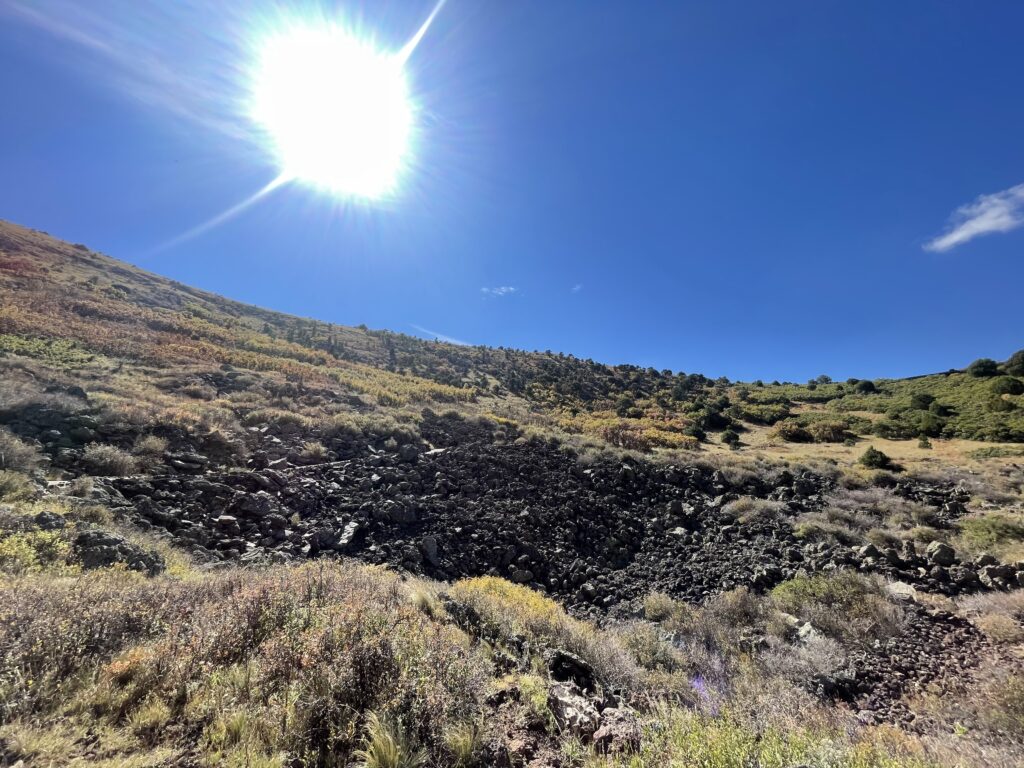 View from the bottom of a crater, a bright sun shines over a sloping hillside with green bushes and brown, dry grass complete with black, lava rocks comprising much of the hillside; all under a clear, blue sky.