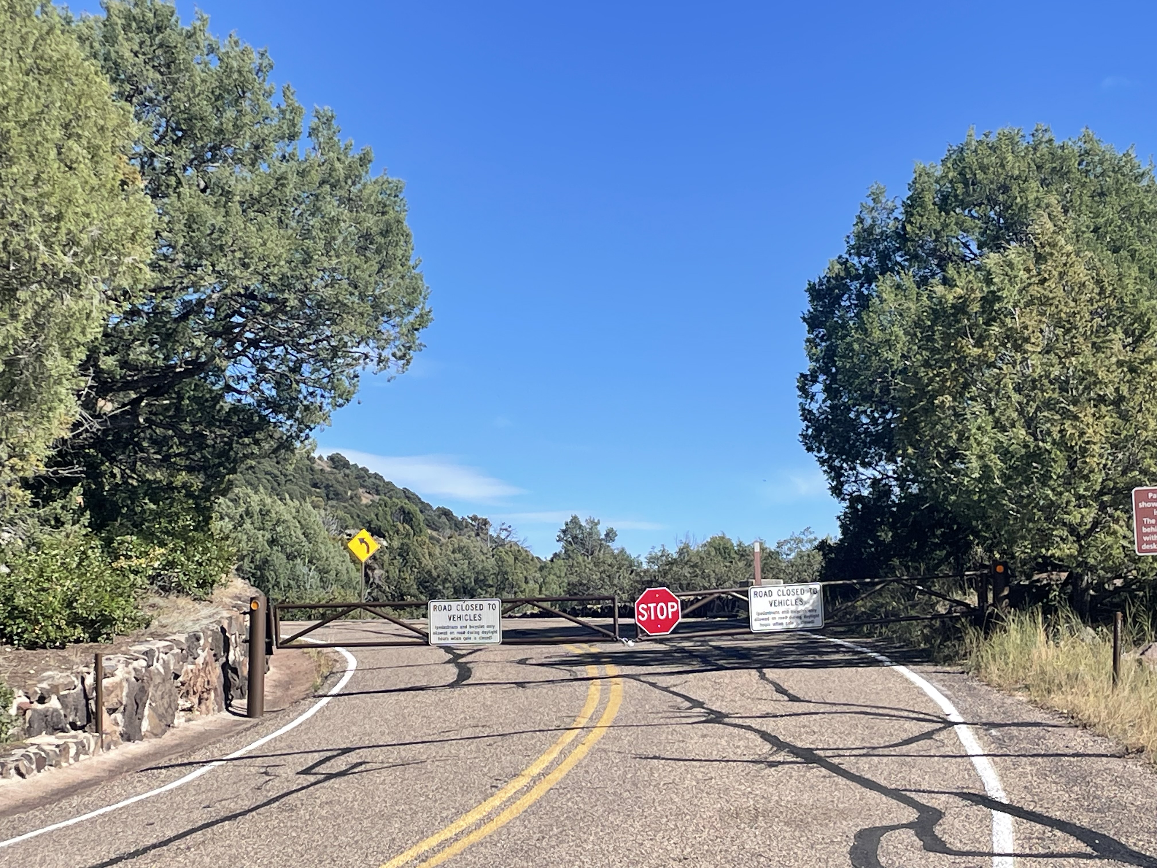 A paved road ends at a closed gate with several warning signs and a stop sign. Trees and greenery line both sides of the road under a clear blue sky.