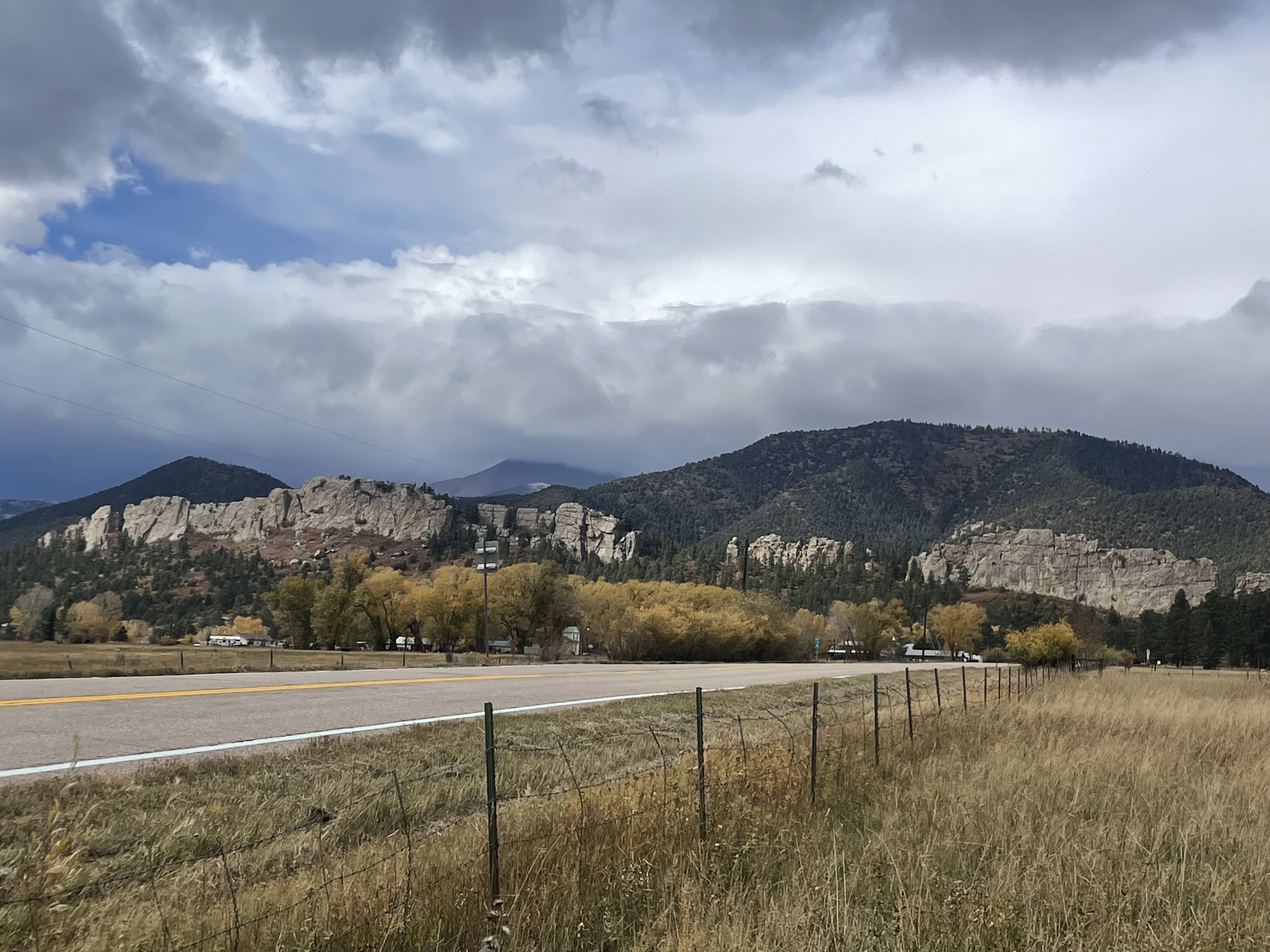 A two-lane road runs past a wire fence and grassy field, with rocky hills, scattered trees, and cloudy skies in the background.