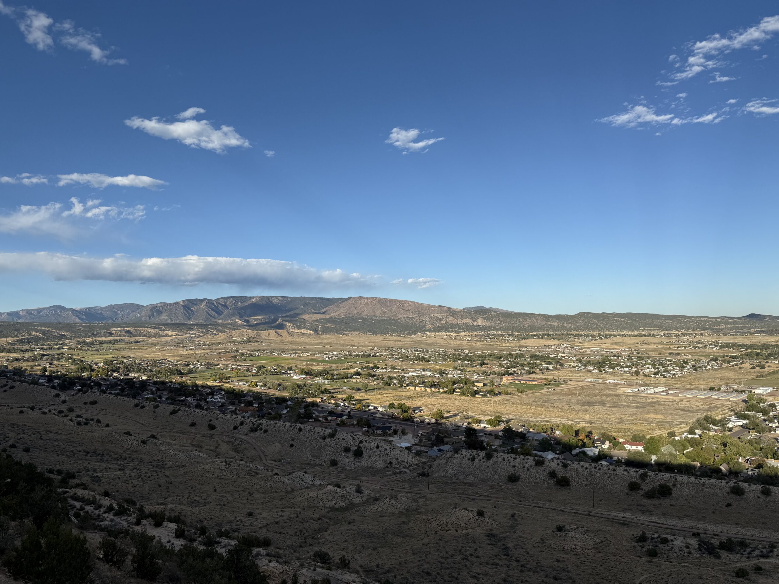 A wide landscape view of a small town with scattered houses, open fields, and mountains in the distance under a clear blue sky with a few clouds. Sunlight casts shadows across the valley.