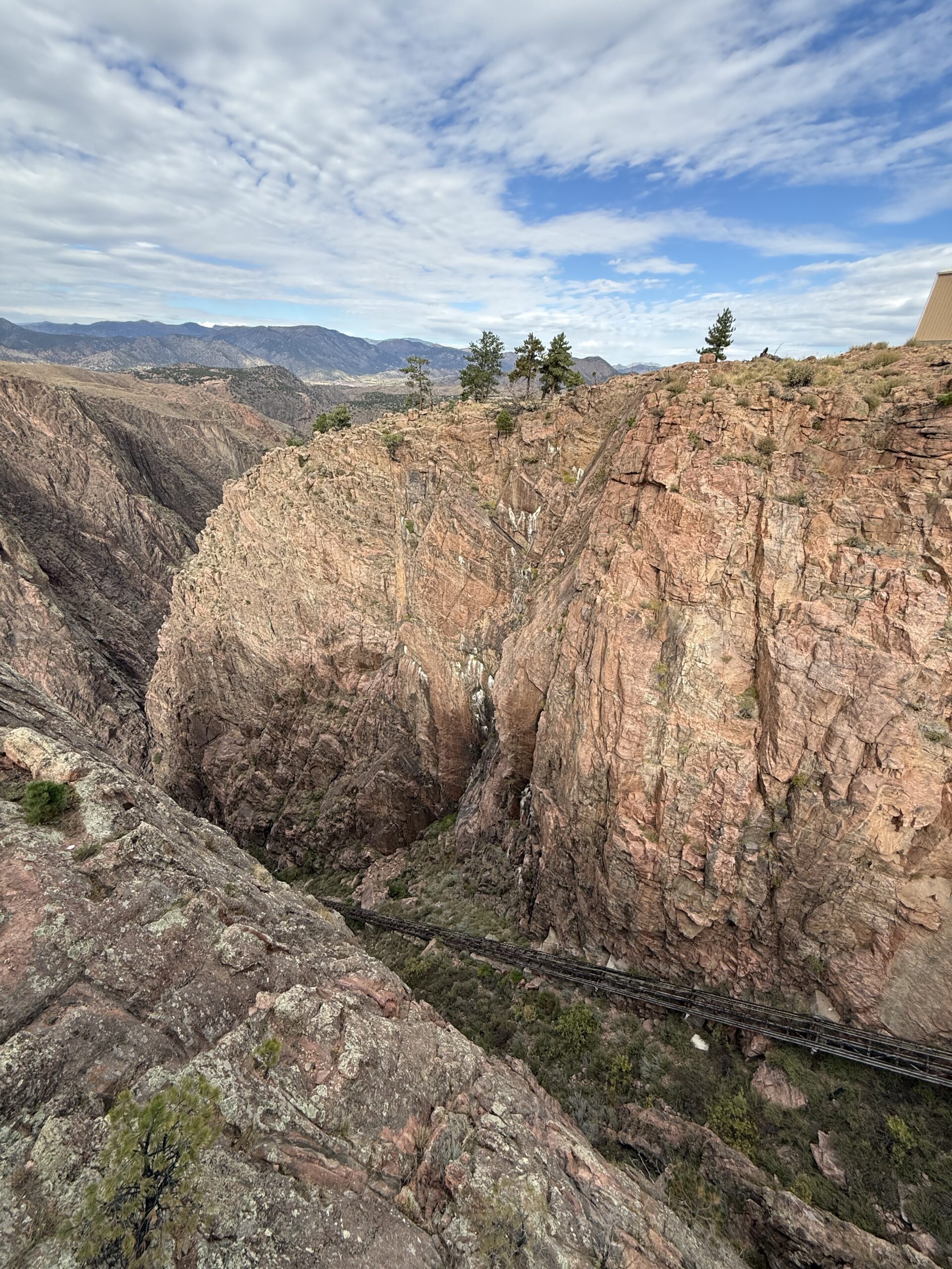 A steep, rocky canyon with reddish cliffs, sparse green trees on top, and a narrow railway track running down the side of the canyon, under a partly cloudy sky. Mountains can be seen in the background.