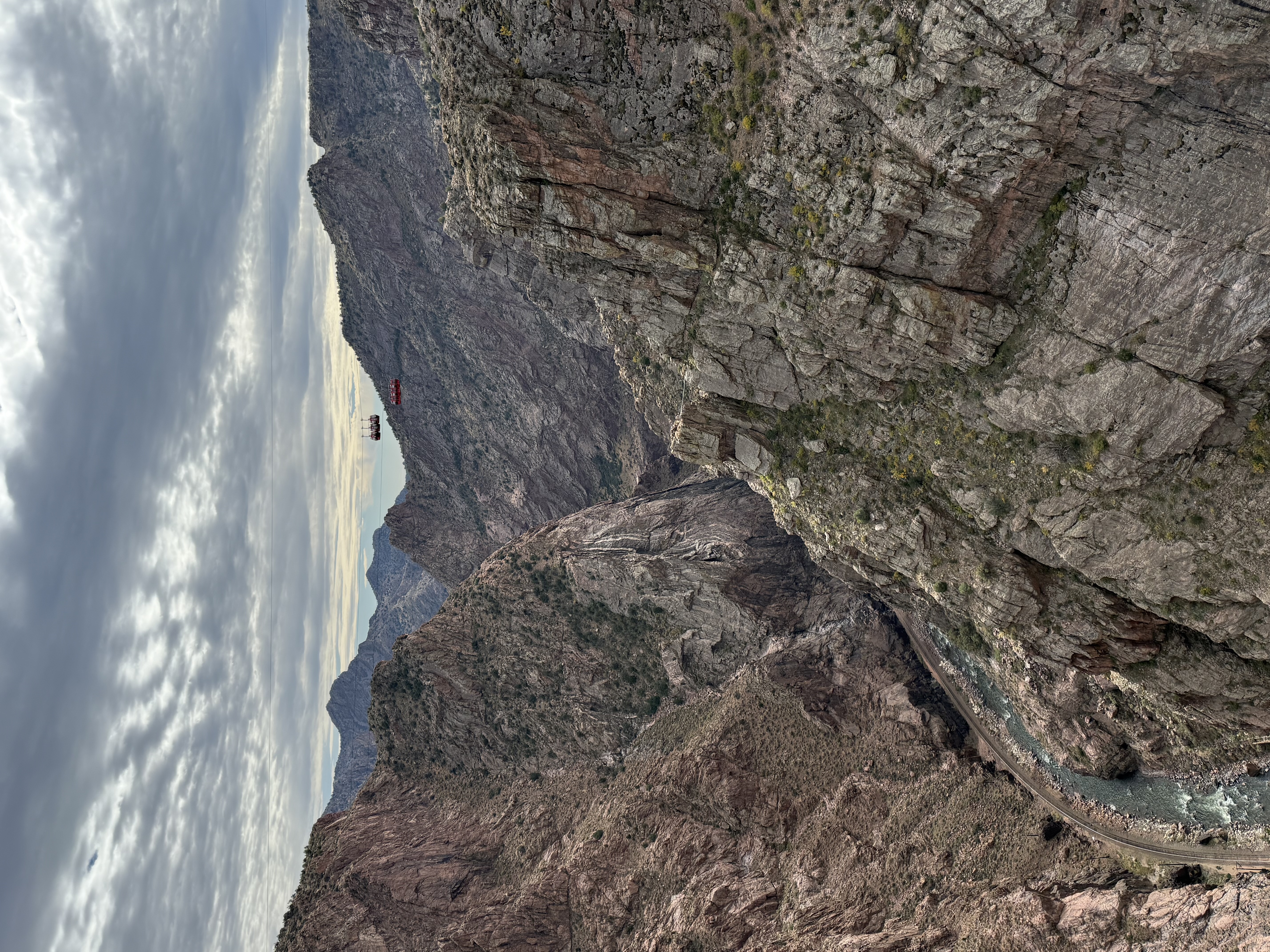A deep rocky canyon with a river flowing at the bottom, surrounded by steep cliffs. A red tramway car travels across the canyon on cables under a cloudy sky.